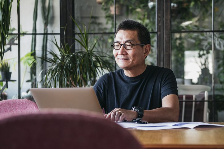 Smiling mature Chinese man achieving deep work on laptop at home