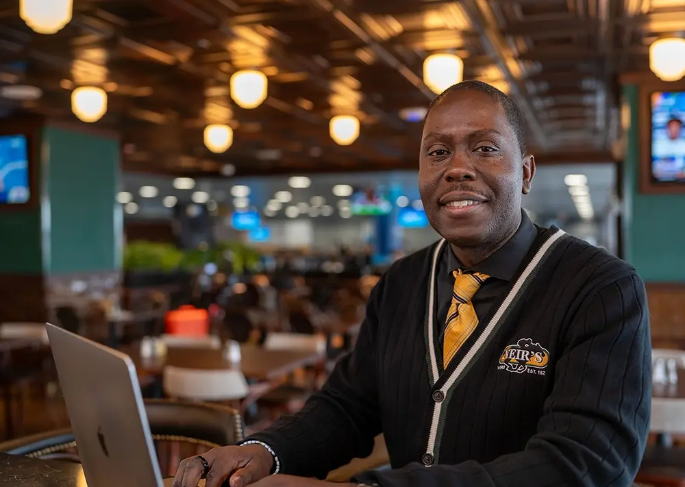 Loy Gordon of Neir's Tavern smiling for the camera sitting at the bar working with his laptop open